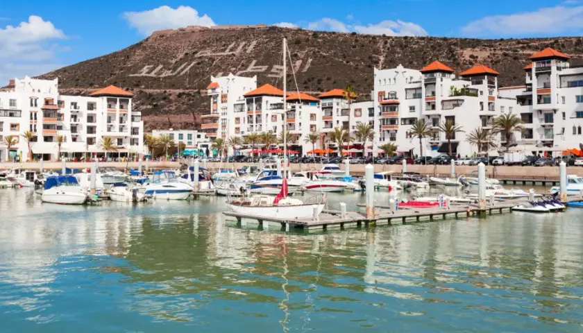 Port de plaisance à Agadir, Maroc, avec des bateaux blancs et des bâtiments modernes. La colline derrière porte une inscription en arabe.