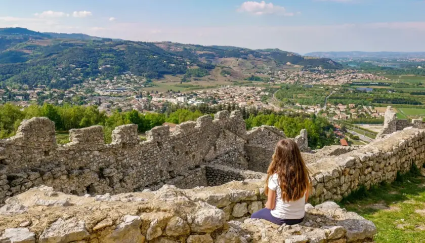 Une jeune fille contemple la vue depuis les ruines du **château de Crussol**, dominant une ville et des collines verdoyantes sous un ciel bleu.
