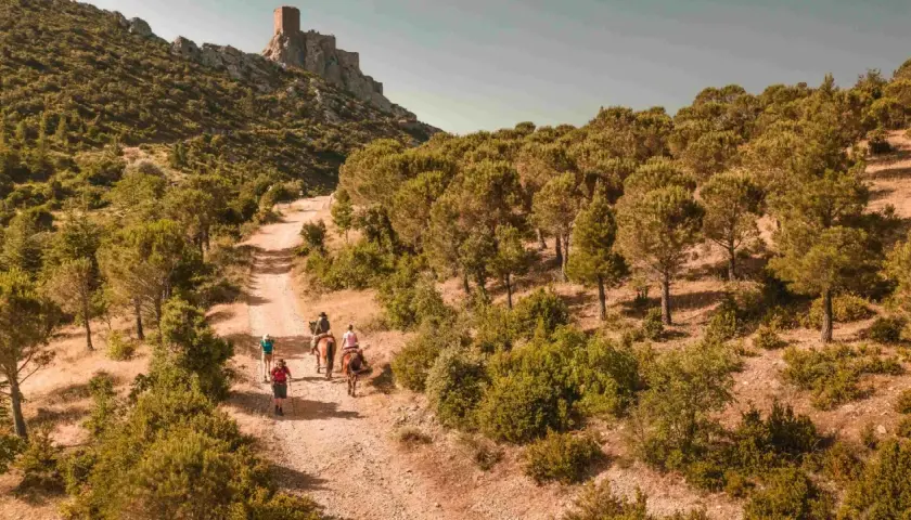 Randonneurs et cavaliers sur le sentier cathare GR 367, menant vers un château perché sur la montagne. Paysage aride et boisé.