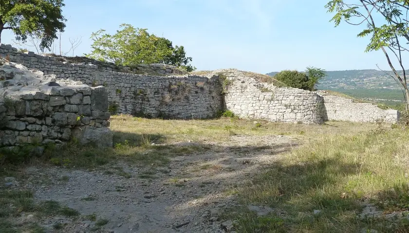 Ruines d'un oppidum gaulois en pierre, avec des murs et des arbres sous un ciel bleu.