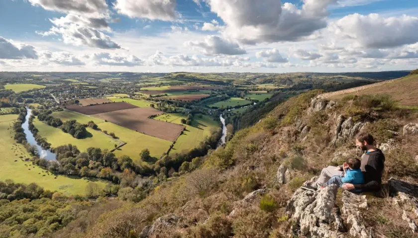 Vue panoramique de la Suisse Normande, une carte vivante de collines verdoyantes, d'une rivière sinueuse et d'un père et son enfant contemplant le paysage.