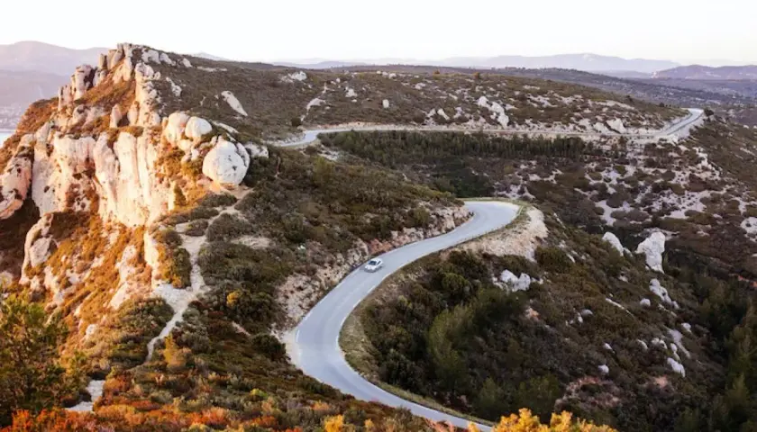 Route sinueuse à travers le massif du Cap Canaille, baignée par la lumière dorée du soleil couchant. Une voiture blanche serpente sur l'asphalte.