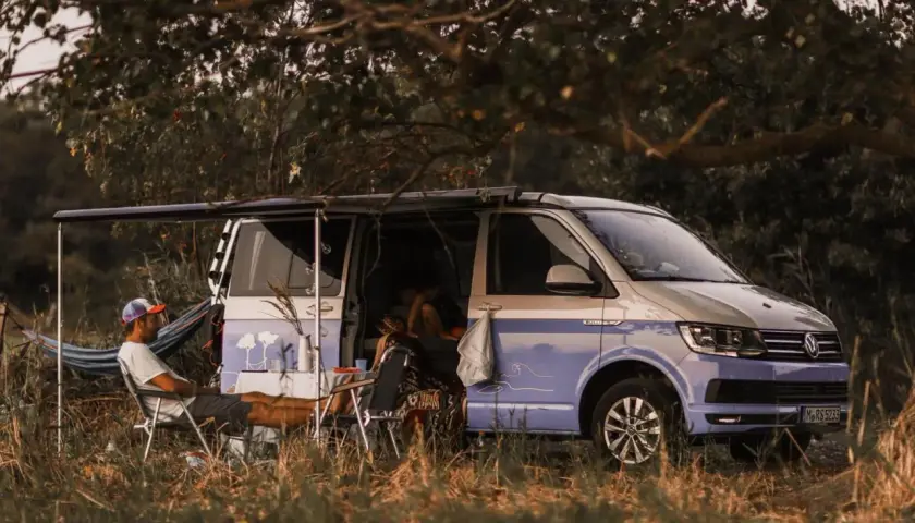 Un homme se détend près d'un camping-car sauvage, avec une table et des chaises installées à l'extérieur. Le véhicule est garé dans un cadre naturel verdoyant.