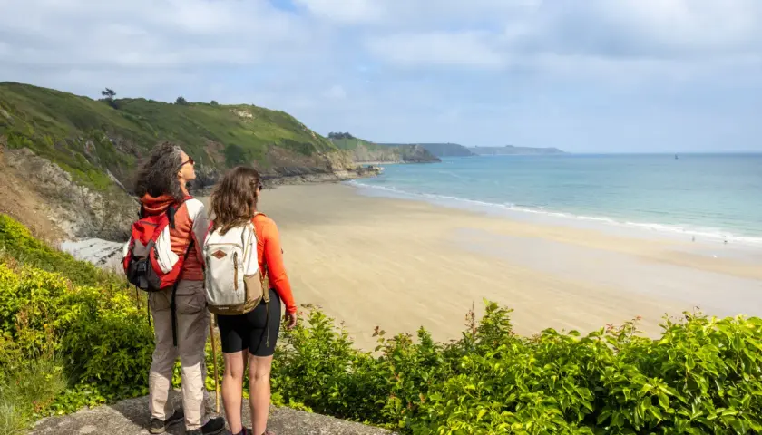 Deux randonneuses contemplent la plage de sable fin et la mer bleue, un paysage typique du GR34 à Saint-Brieuc.