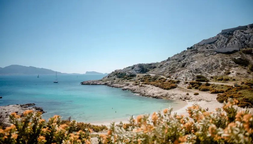 Vue panoramique d'une crique aux eaux turquoise sur la plage frioul, avec des voiliers au loin et des collines rocheuses.