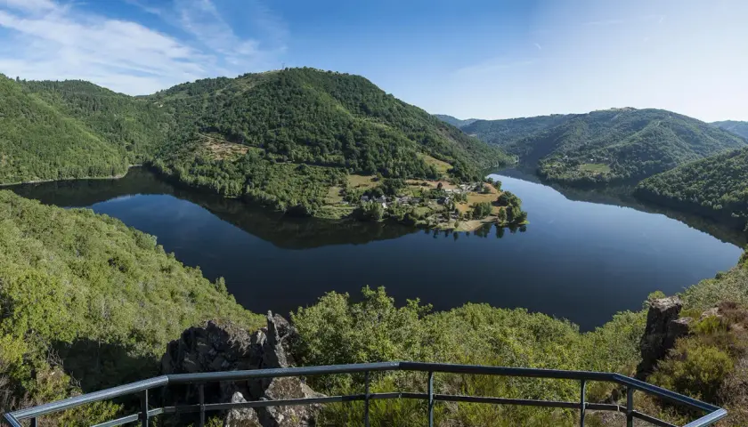 Vue panoramique des gorges de la Truyère, avec un lac paisible reflétant les collines verdoyantes et un village pittoresque.