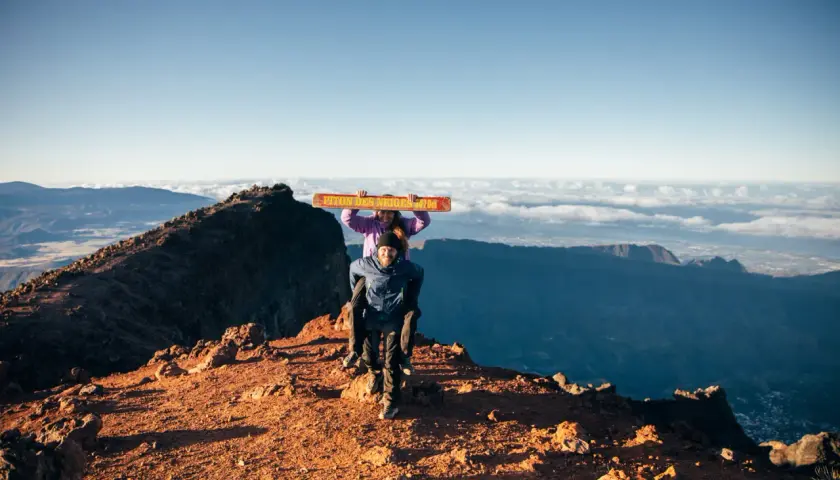 Couple joyeux au sommet du Piton des Neiges, portant une pancarte "Piton des Neiges 3070m". Paysage spectaculaire au lever du soleil.