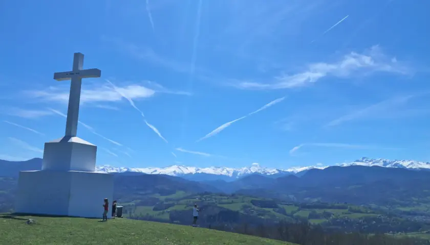 Le tuc de Montcalivert domine un paysage verdoyant sous un ciel bleu zébré de traînées de condensation. Une grande croix blanche se dresse fièrement.