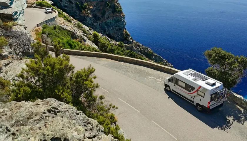 Un camping-car blanc circule sur une route côtière sinueuse en Corse, dominant la mer d'un bleu profond.