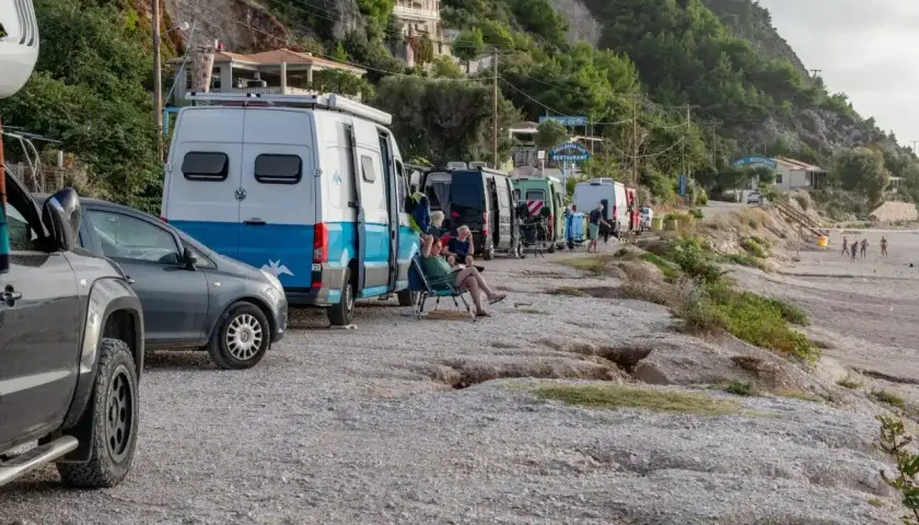 Une rangée de camping-cars en Grèce, certains garés sur un chemin de gravier près d'une plage. Des gens se détendent à l'extérieur.