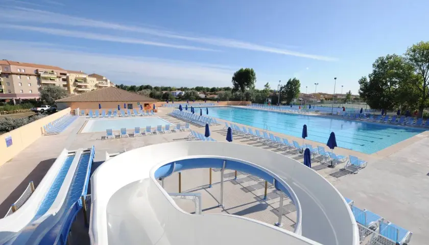 Piscine et toboggans aquatiques dans une aire de camping-car au Grau-du-Roi. Des chaises longues bleues et des parasols sont disposés autour des bassins.