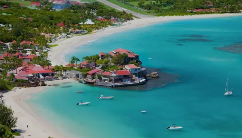 Vue aérienne de Saint-Barthélemy, avec ses plages de sable blanc, ses eaux turquoise et ses villas aux toits rouges. Un voilier navigue paisiblement.