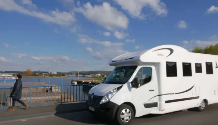 Un camping-car blanc roule sur un pont, avec une personne marchant à côté. Le ciel est bleu avec des nuages. Idéal pour la consommation camping-car.
