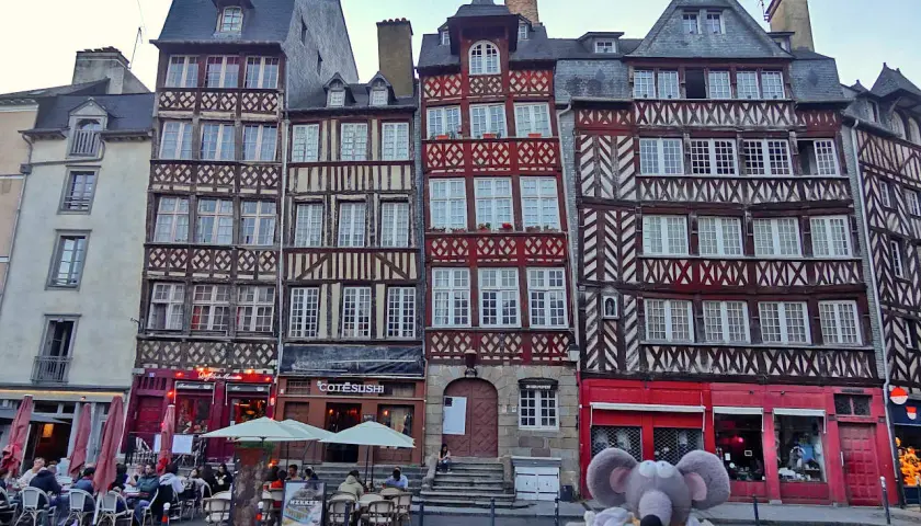 Une souris en peluche pose devant des maisons à colombages colorées, un aperçu de ce que faire à Rennes.