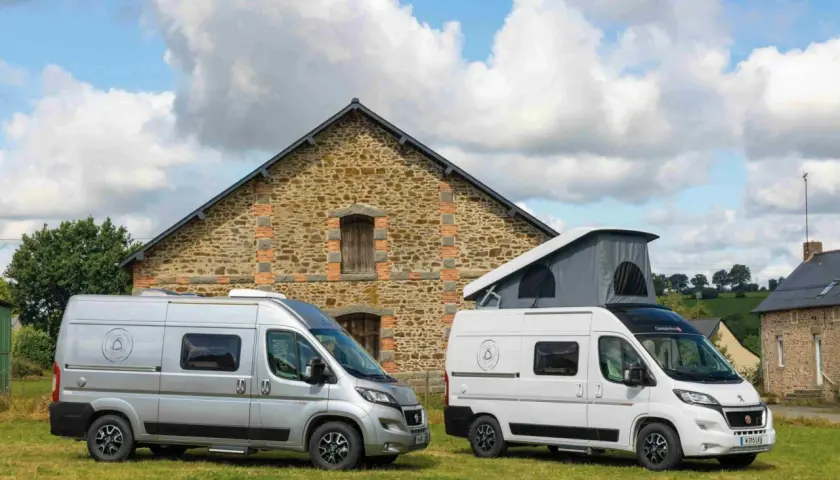 Deux camping-cars, un argenté et un blanc avec toit relevable, devant une bâtisse en pierre. Idéal pour savoir comment choisir son camping-car.