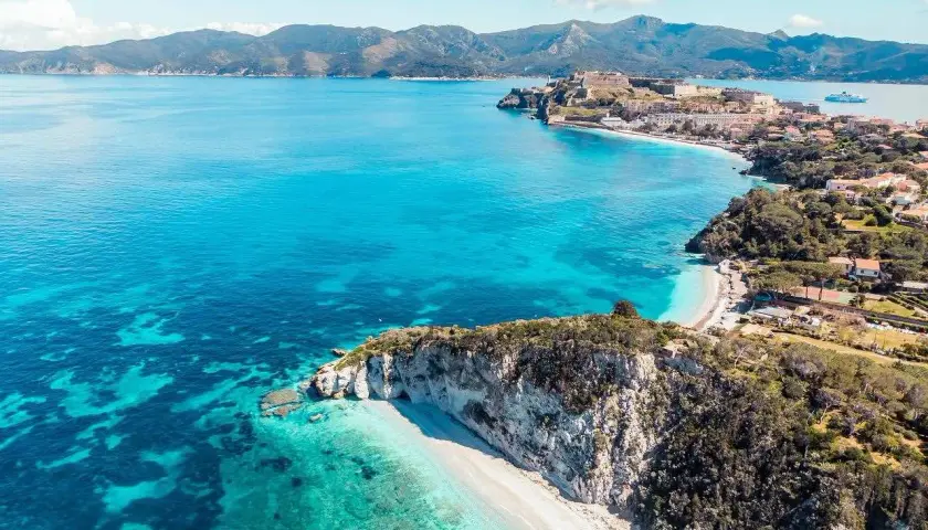 Vue aérienne de l'île d'Elbe : falaises, plages de sable blanc, eaux turquoise et ville fortifiée. Que faire ? Explorer, nager, se détendre.