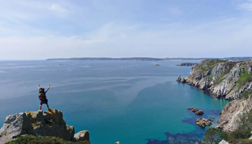 Randonneur victorieux au sommet d'un rocher, bras levés, dominant la mer turquoise et les falaises de GR34 Crozon.