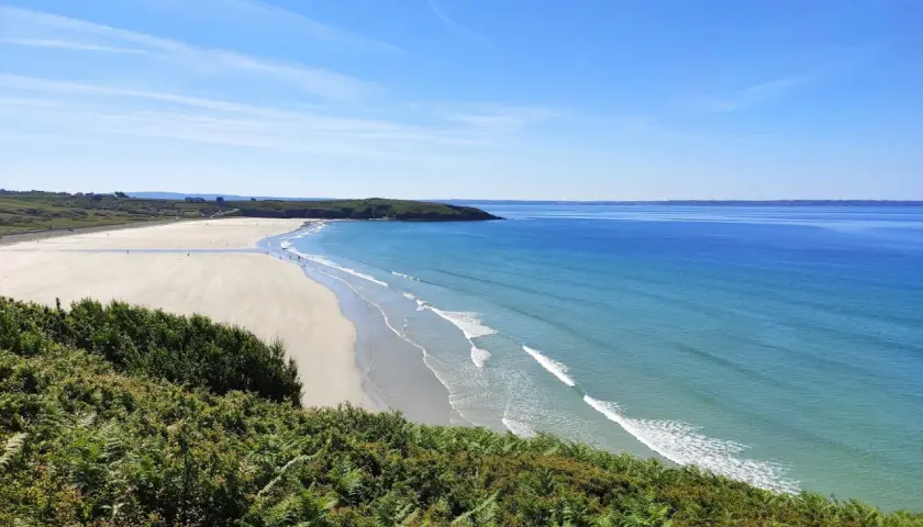 Vue panoramique d'une plage trez bellec, avec sable clair, mer bleue et végétation verdoyante au premier plan.