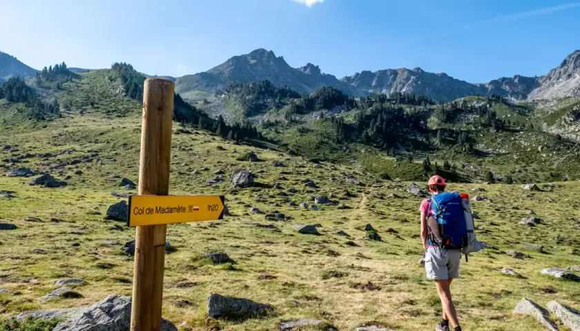 Randonneuse sur le GR10, suivant le panneau jaune indiquant le Col de Madamète. Paysage de montagne verdoyant.