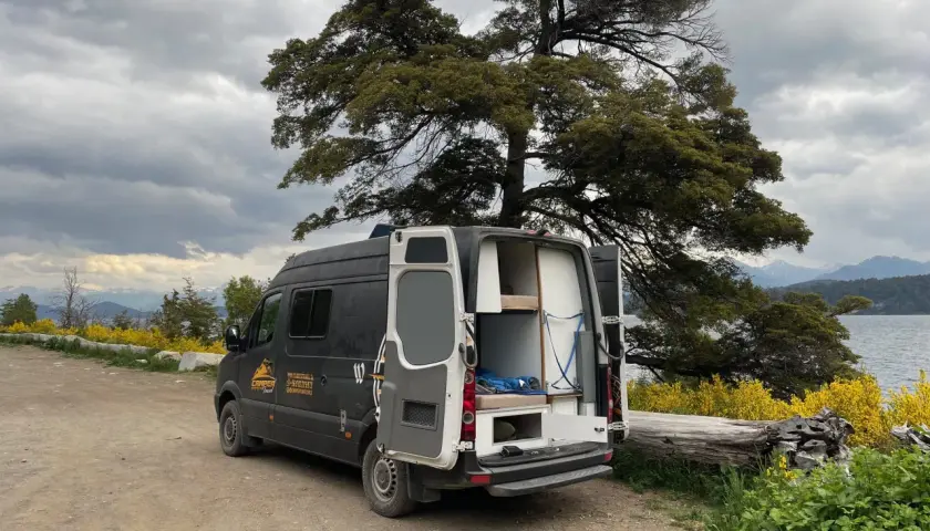 Un camping-car noir avec les portes arrière ouvertes, prêt pour un entretien, est garé près d'un lac sous un ciel nuageux.