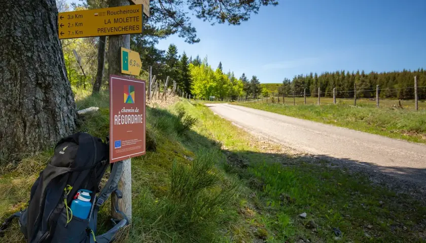 Sac à dos prêt pour l'aventure sur le chemin de Regordane. Panneaux indicateurs et route s'étendent sous un ciel bleu.