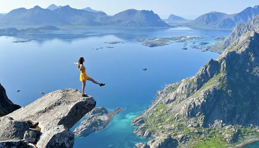 Une femme en robe jaune sur un promontoire rocheux, admirant les plus beaux endroits des îles Lofoten, avec des montagnes bleues et des eaux turquoise.