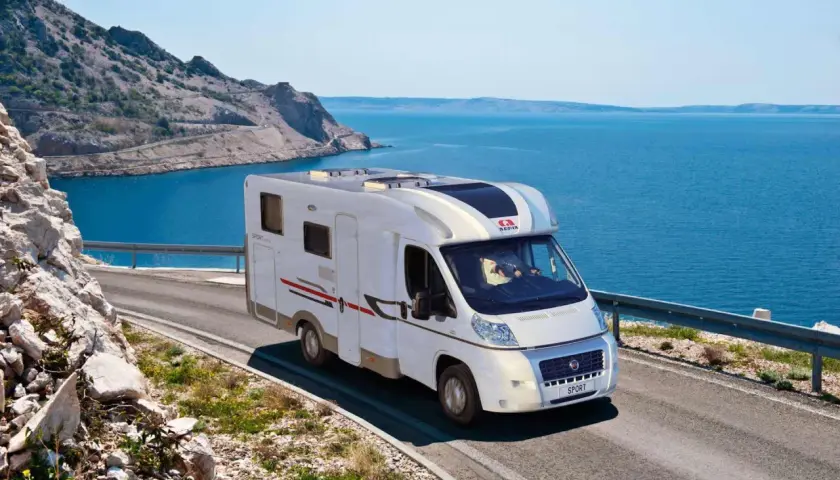 Un camping-car Adria roule sur une route côtière en Sardaigne, avec la mer bleue et des montagnes rocheuses en arrière-plan.