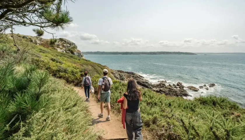 Trois randonneurs explorent le sentier des douaniers, longeant la côte rocheuse et la mer d'un bleu profond sous un ciel nuageux.