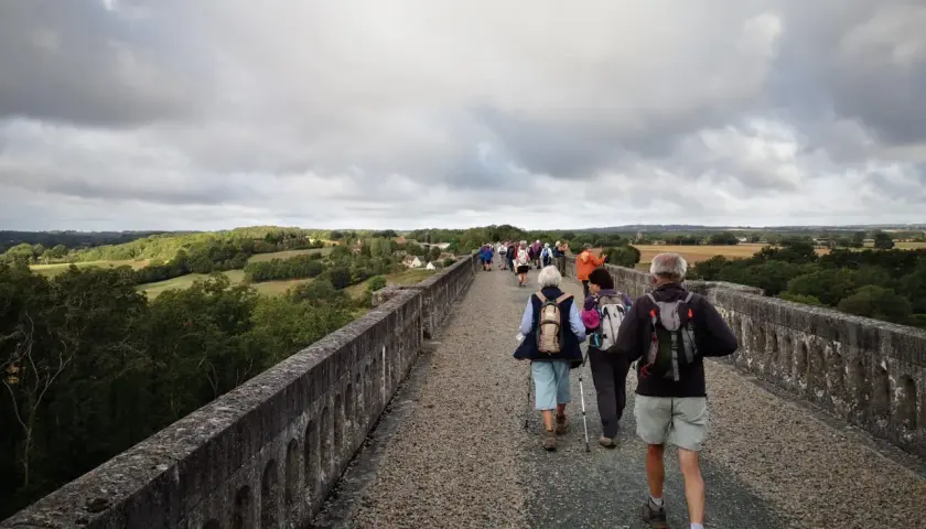 Un groupe de randonneurs, dont certains portent des sacs à dos et utilisent des bâtons de marche, traverse un ancien aqueduc en pierre sous un ciel couvert. Le paysage verdoyant s'étend à perte de vue.