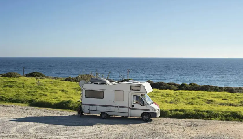 Camping-car blanc garé sur une aire gratuite en bord de mer en Bretagne. L'océan bleu s'étend à l'horizon, avec des champs verdoyants.