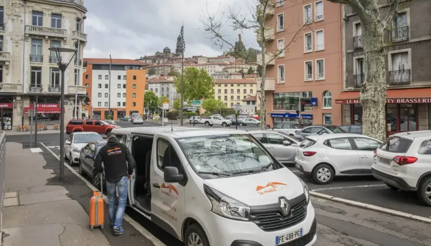 Un homme charge une valise orange dans un bus compostelle blanc. La ville s'étend en arrière-plan.