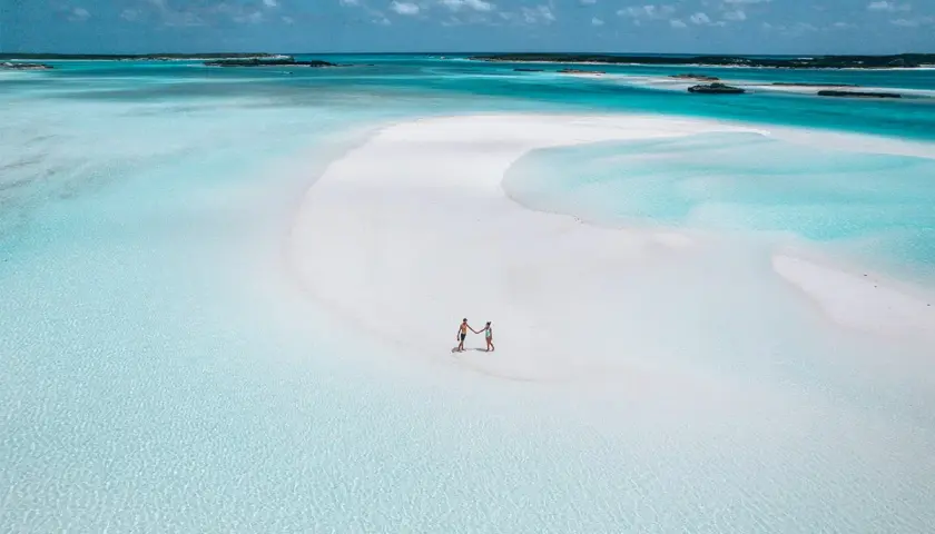 Un couple se tient la main sur une plage de sable blanc aux Bahamas. L'eau turquoise et les îlots au loin invitent à explorer que faire aux Bahamas.