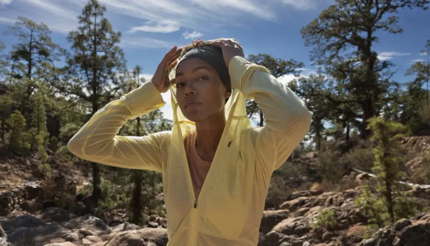 Une femme ajuste son bandeau et sa veste légère, parfaite pour savoir comment s'habiller pour une randonnée en été. Paysage rocheux et arbres en arrière-plan.