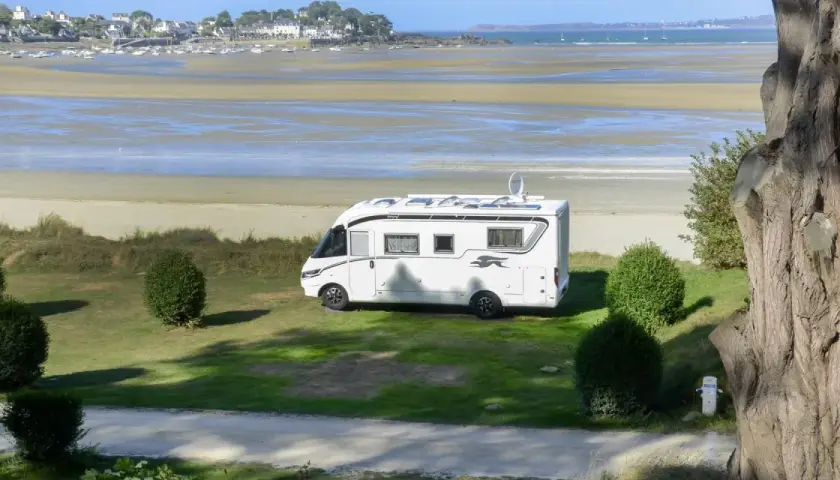 Vue d'une aire de camping-car avec sanitaire, face à une plage et un village côtier. Le camping-car blanc est garé sur l'herbe.