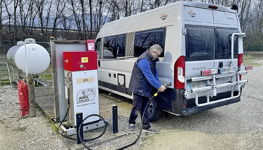 Un homme fait le plein de son camping-car à une installation GPL. Le véhicule est garé à côté d'une pompe à carburant rouge et blanche.