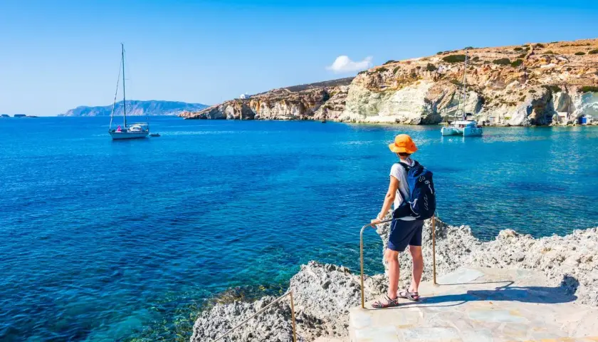 Un voyageur admire la mer Égée bleue et les falaises blanches, un voilier au loin. Le tourisme en Grèce offre des paysages magnifiques.