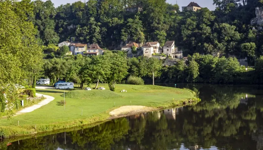 Vue paisible d'un camping-car près de la rivière, avec des maisons pittoresques accrochées à la colline, rappelant le charme de Figeac et Cahors.
