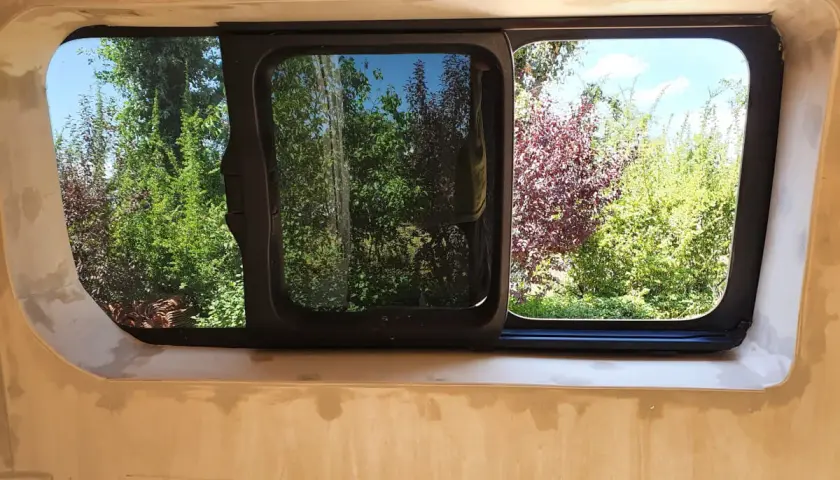 Fenêtre de fourgon aménagé, prête pour le contrôle technique. Vue sur un jardin verdoyant et fleuri.