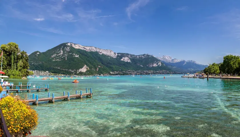 Vue panoramique du lac d'Annecy, avec des montagnes verdoyantes en arrière-plan. Des bateaux et des pédalos animent les eaux turquoise. Un ponton fleuri invite à la promenade.