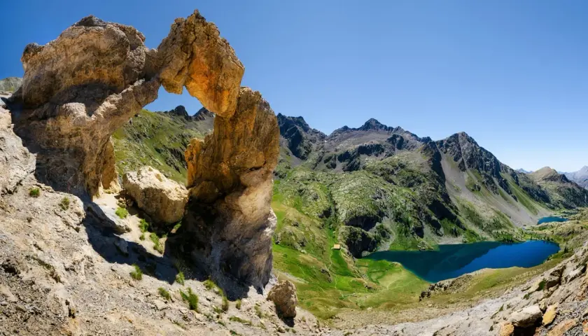 Un arc rocheux spectaculaire domine un paysage de montagne verdoyant, avec des lacs bleus scintillants. Une vue digne d'une carte du Mercantour.
