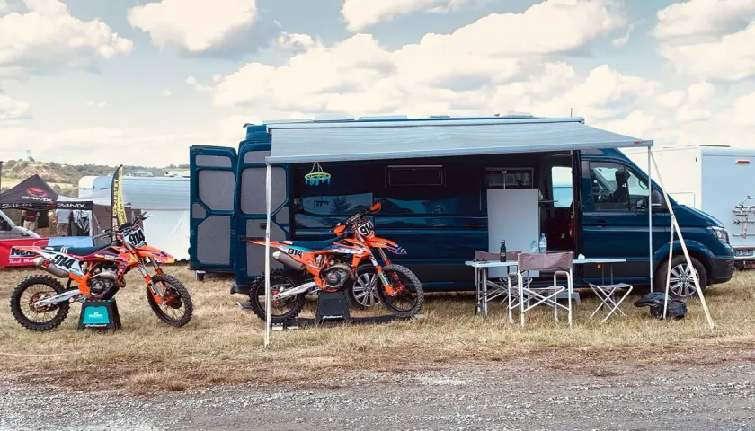 Un fourgon aménagé avec soute moto, prêt pour l'aventure. Deux motos de cross orange attendent devant, sous un ciel nuageux.