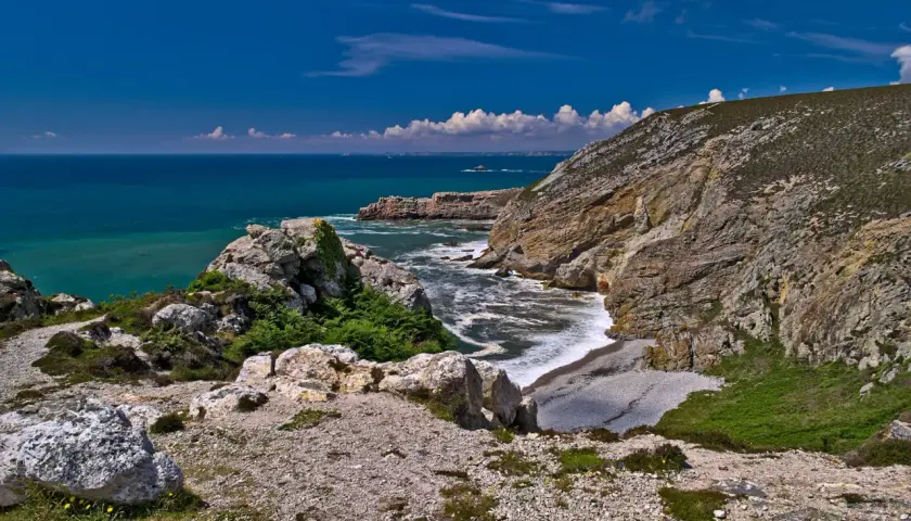 Sentier rocailleux menant au **Cap de la Chèvre**, avec falaises abruptes plongeant dans une mer turquoise et un ciel bleu éclatant.