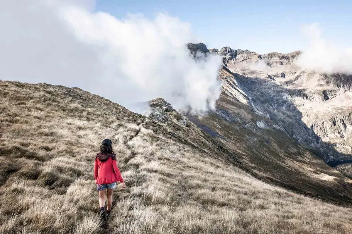 Une randonneuse explore un sentier herbeux, s'approchant des majestueuses montagnes du tour du Biros, enveloppées de nuages.