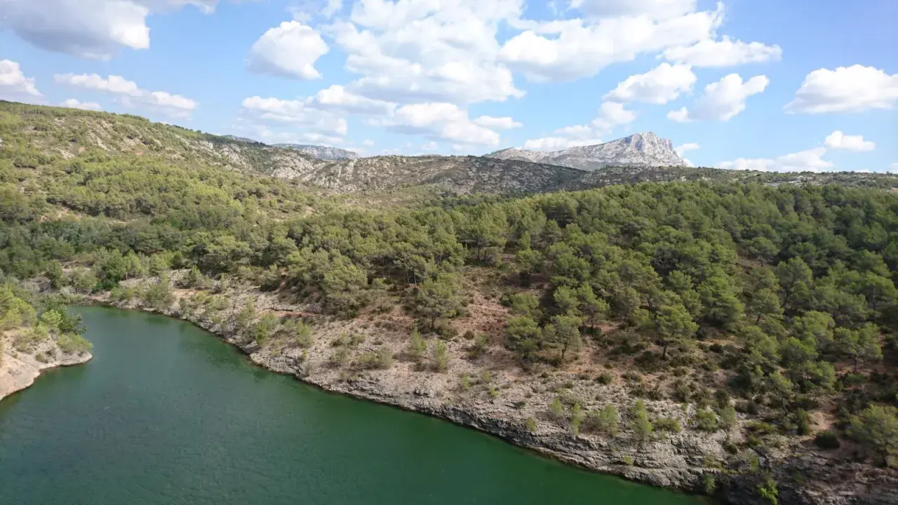 Vue panoramique du lac Zola, entouré de collines verdoyantes et rocheuses sous un ciel bleu parsemé de nuages blancs.