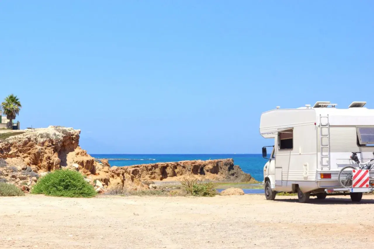 Camping-car blanc stationné sur une plage de sable en Espagne, avec la mer bleue et des falaises rocheuses en arrière-plan. Une aire de camping-car espagne gratuite idéale pour les vacances.