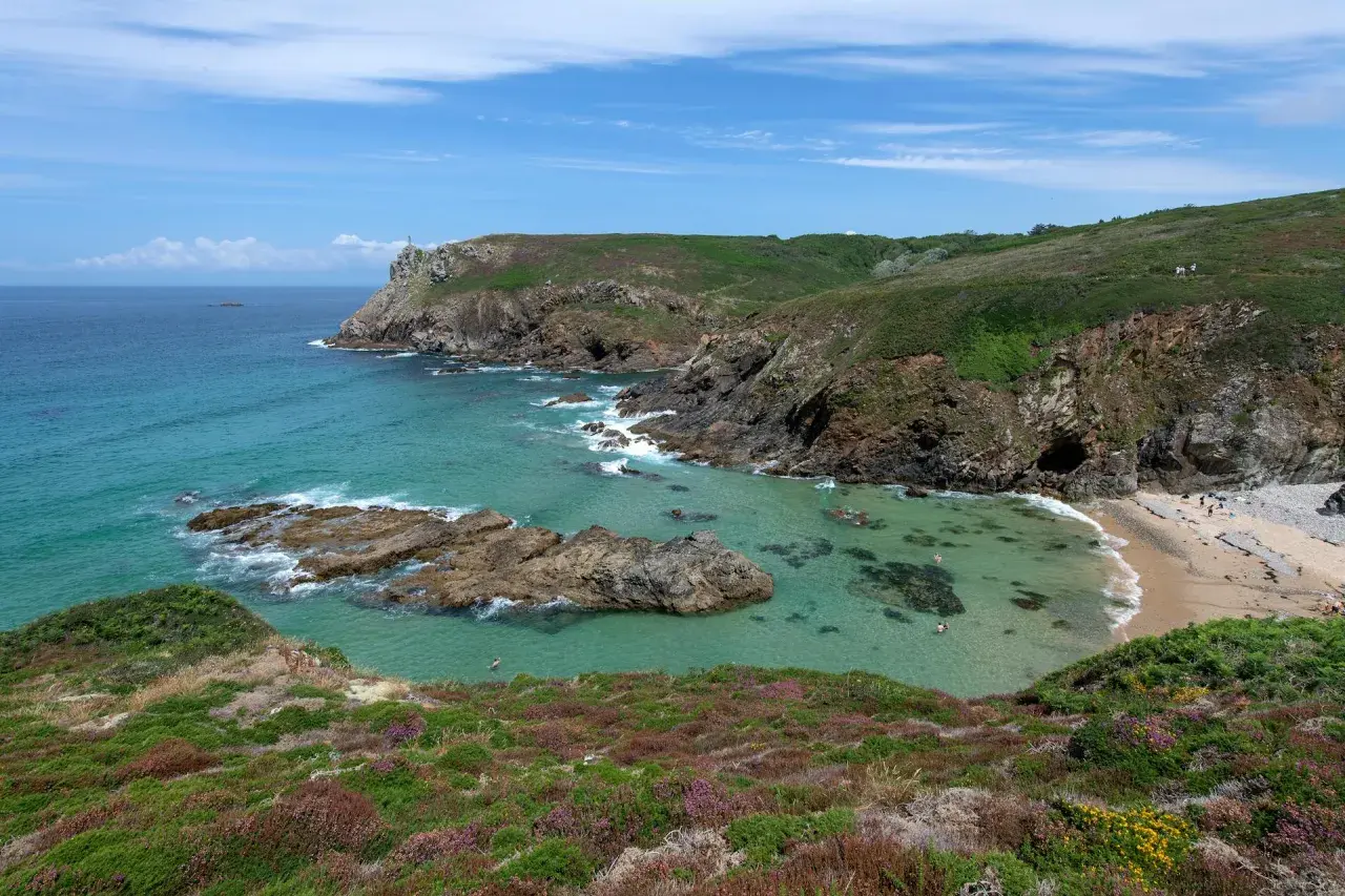 Vue panoramique de la pointe du Van, avec ses falaises verdoyantes, ses eaux turquoise et sa petite plage. Quelques baigneurs profitent du soleil.