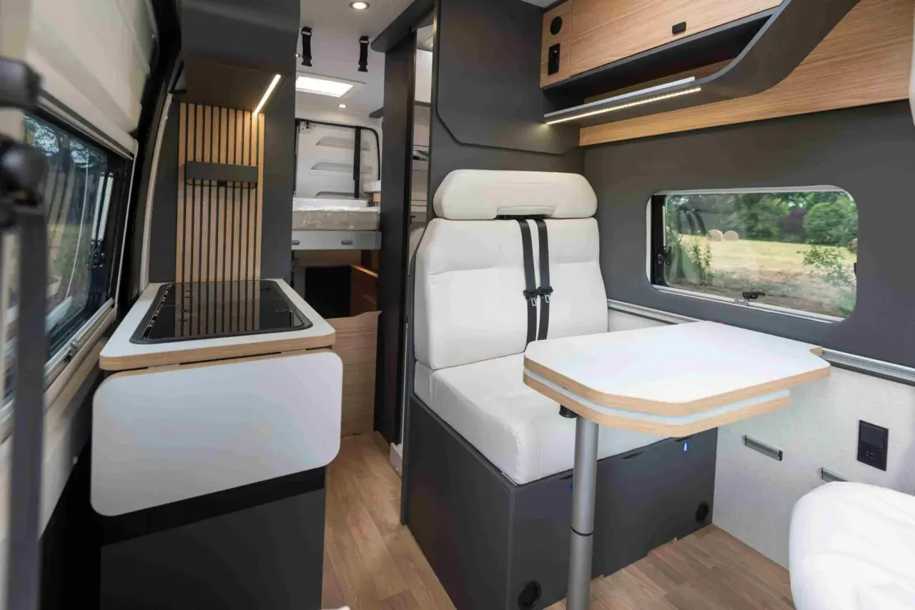 Intérieur d'un camping-car Bavaria : banquette confortable, table escamotable, plaque de cuisson et lit visible au fond.