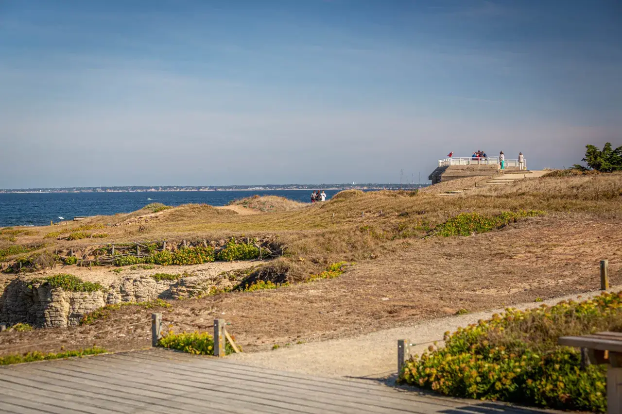Vue panoramique sur la côte sauvage de la pointe Saint-Gildas, avec des falaises rocheuses, des dunes et la mer bleue. Des promeneurs profitent du paysage.