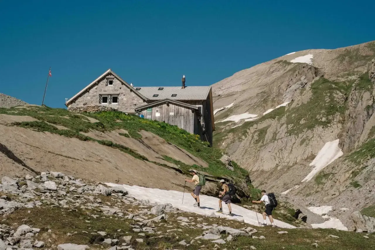 Trois randonneurs approchent d'une **cabane refuge** en pierre et bois, perchée sur une pente rocheuse et herbeuse, sous un ciel bleu intense.