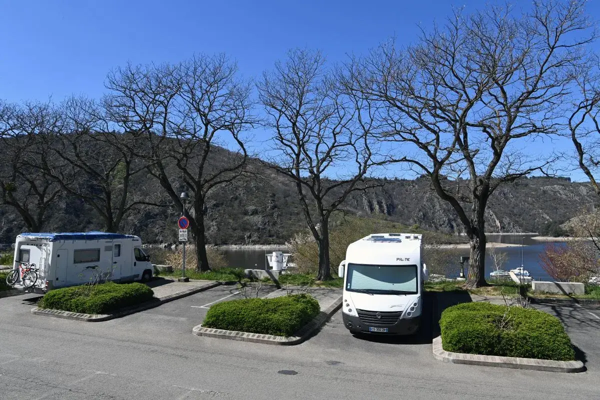 Stationnement pour camping-cars gratuit en Auvergne, avec vue sur le lac et les montagnes. Deux camping-cars sont garés sous des arbres dénudés.